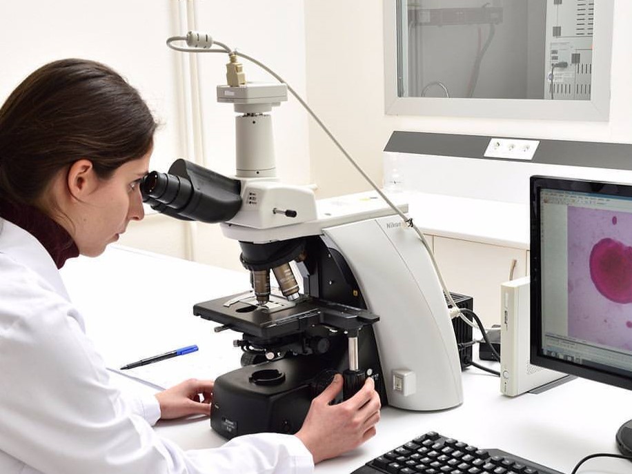 Lab researcher Bee&You in a white coat using a microscope, with a nearby monitor displaying a magnified pink sample.